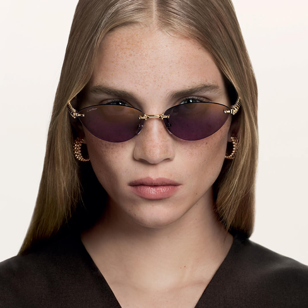 Woman wearing sunglasses with gold frames and purple lenses on a plain background